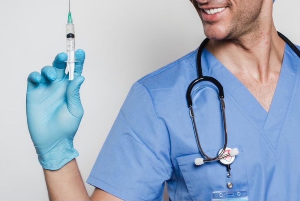 Smiling male medical professional in scrubs and gloves holding a syringe, prepared for an injectable treatment or procedure.