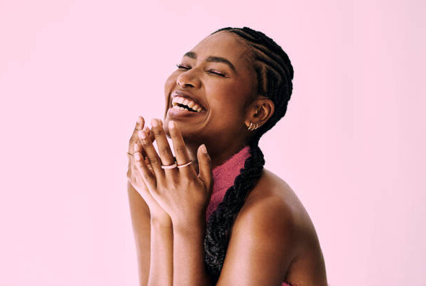 Joyful woman with glowing skin laughing against a pink background, showcasing radiant, healthy complexion.